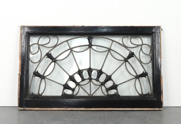 Antique fanlight transom window with a black-painted wood frame, radiating leaded glass design, and raised floral details, shown with mounting hardware and a visible crack in the glass.