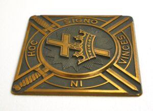 Antique brass and black square plaque with a cross, crown, and diagonal swords, with “IN HOC SIGNO VINCES” lettering and light wear visible on the surface.