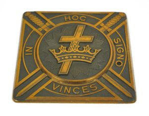 Antique brass and black plaque with a cross and crown emblem, the words “In Hoc Signo Vinces,” and diagonal swords in a square design.