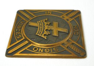 Antique brass and black square plaque with a cross, crown, and diagonal swords, showing the words “In Hoc Signo Vinces” and light wear.