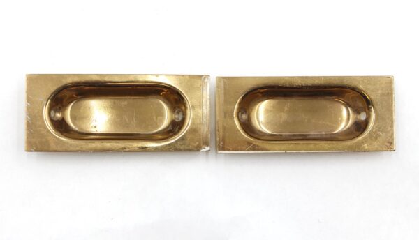 Pair of polished brass sash lifts shown side by side, each with an oval recessed pull and mounting holes on a rectangular backplate.