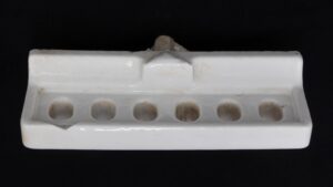 White ceramic wall-mount toothbrush holder with six holes, shown front on with visible surface wear and a chip at the left front edge.