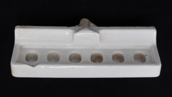 White ceramic wall-mount toothbrush holder with six holes, shown front on with visible surface wear and a chip at the left front edge.
