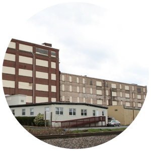 An industrial brown and beige building with many windows stands behind a small white building by train tracks in Scranton, Pennsylvania, all under a cloudy sky.
