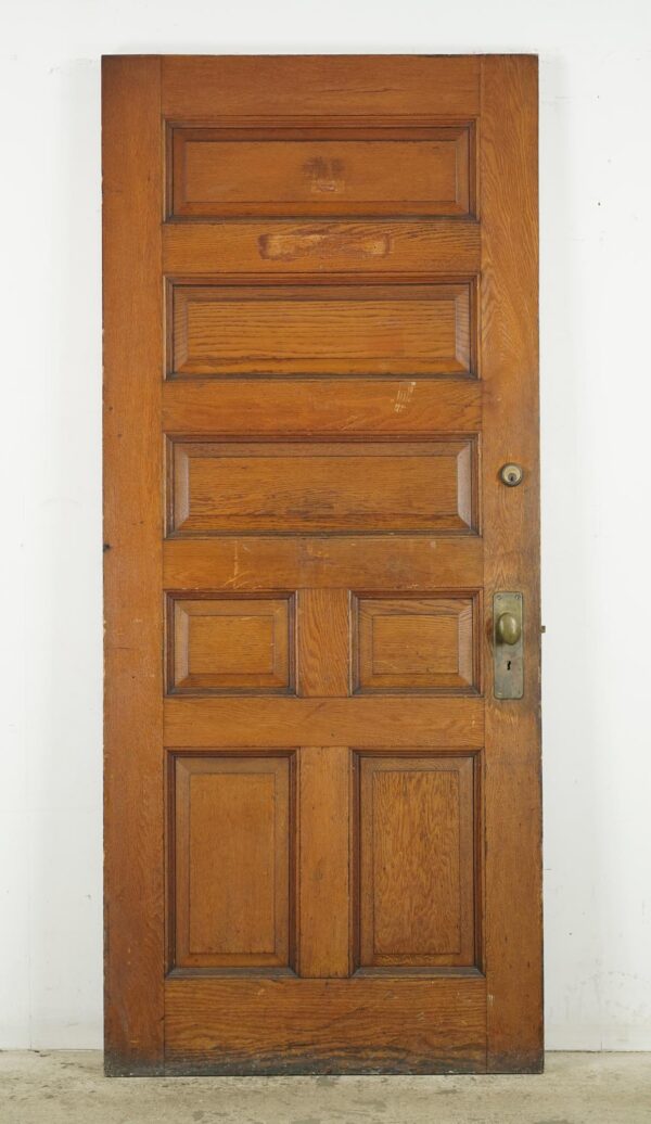 Antique chestnut entry door with seven panels and attached brass hardware, shown full length with visible surface wear.