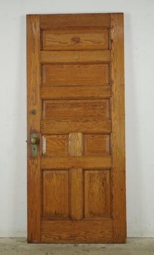 Antique chestnut entry door with seven panels and attached brass and steel hardware, shown front view with visible surface wear and a slanted worn bottom edge.