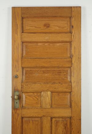 Antique seven-panel entry door in a medium wood tone, shown front view with some original brass hardware attached and visible surface wear.