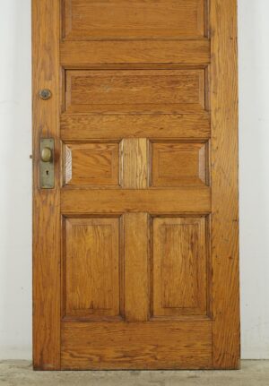 Antique seven-panel entry door in a medium wood tone with original brass hardware attached, shown front-facing with visible surface wear and a slightly worn bottom edge.