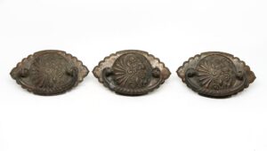 Three views of a Victorian brass drawer bail pull with an oval backplate, scalloped edges, and ornate scrollwork, shown with an antique patina and age wear.