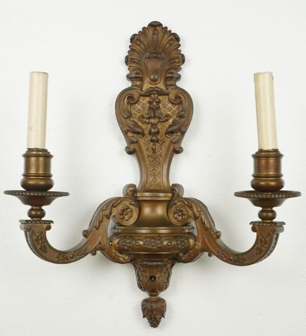 Ornate bronze two-arm wall sconce with classical scrollwork, acanthus leaves, and two candle sockets.