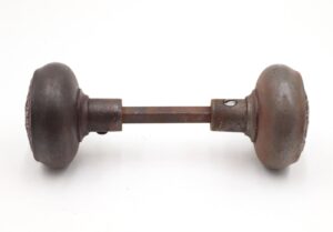 Pair of cast iron passage door knobs with a spindle, shown side by side and connected in the center, with visible surface wear and a dark finish.