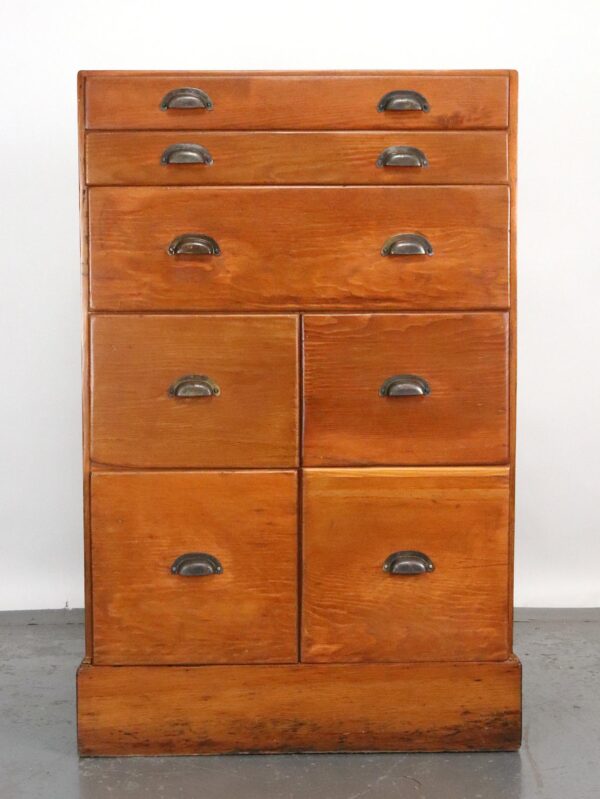 Front view of an antique pine apothecary cabinet with seven drawers and black cast iron cup pulls, shown in a medium wood tone with visible surface wear near the base.
