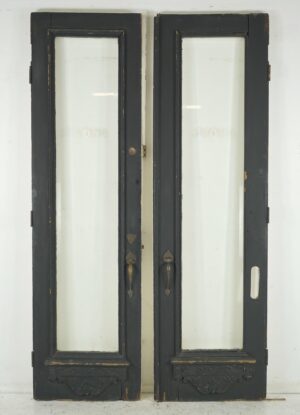Vintage black wood double doors with tall glass panes, brass hardware, and ornate carved trim, shown closed with surface wear visible.