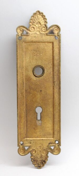 Neoclassical bronze entry back plate with ornate scroll and floral ends, a central keyhole, and four mounting holes.