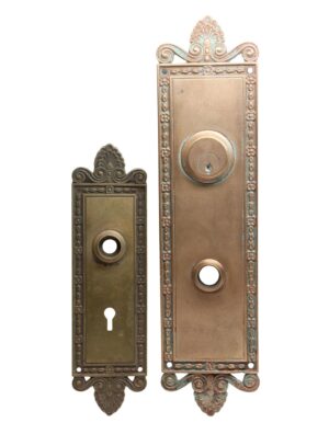 Pair of neoclassical bronze door back plates shown side by side, one taller entry plate and one shorter interior plate, both with ornate scroll and floral detailing and keyhole openings.