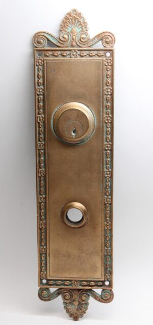 Neoclassical bronze entry back plate with ornate scroll and floral details, a round lock opening, and an antique bronze patina.