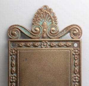 Neoclassical bronze entry back plate with an ornate scroll-and-fan crest, rectangular center, and two mounting holes visible at the top corners.