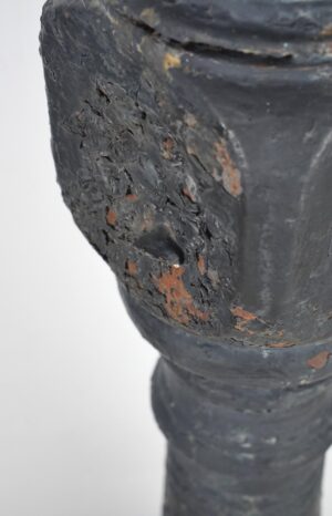 Close-up of a black cast iron newel post with chipped paint and rust, showing a worn embossed rosette detail and part of the rounded finial.