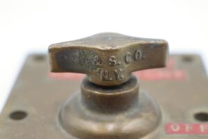 Close-up of a brass ship light switch with a T-shaped knob stamped “R & S CO NY.”