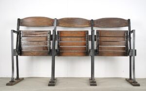 Three-seater folding stadium seat unit with slatted wood seats and backrests on a black steel frame, shown folded upright with four floor supports.