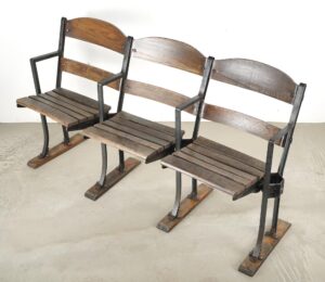 Three-seater folding stadium seat with slatted wood backs and seats on a black steel frame, shown at an angle with visible bottom stabilizing feet and minor wear in the wood and metal.