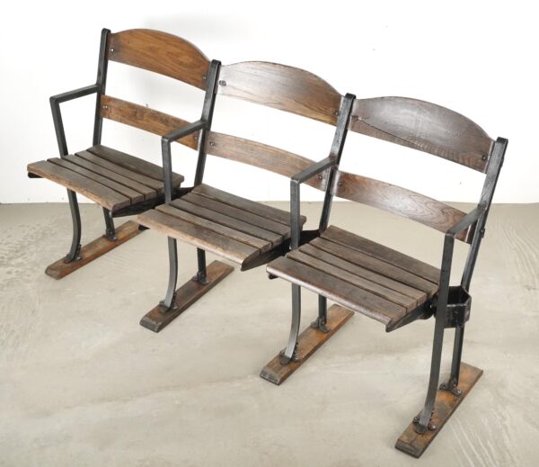 Three-seater folding stadium seat with slatted wood backs and seats on a black steel frame, shown at an angle with visible bottom stabilizing feet and minor wear in the wood and metal.