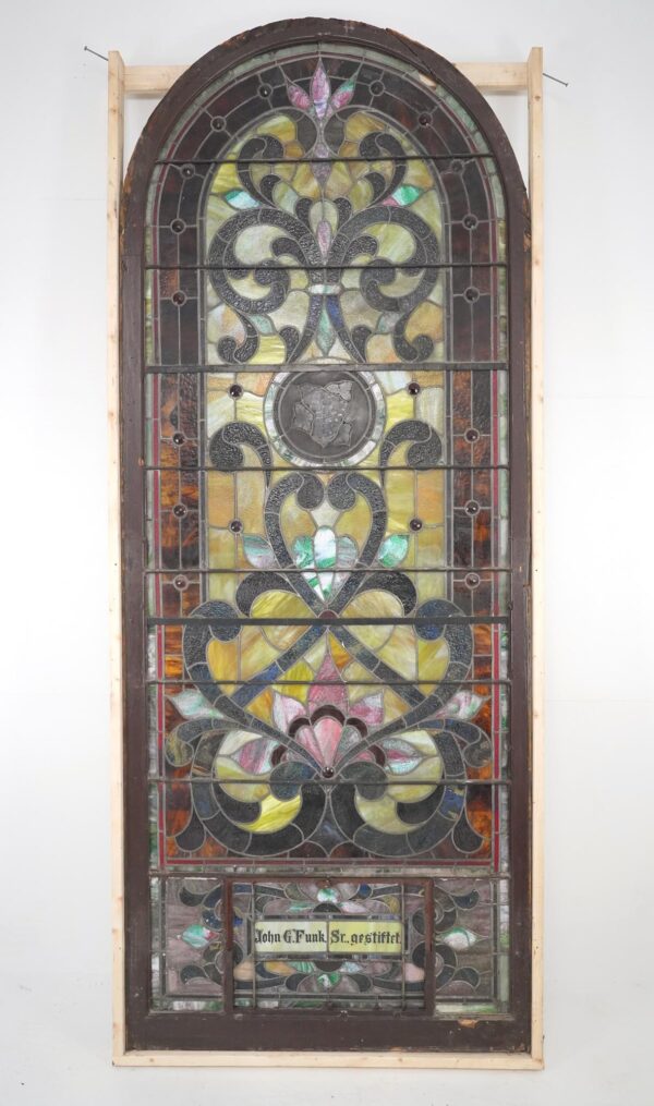 Tall arched stained glass church window with multicolored scrolling vine and leaf panels, brown frame, and a small name plaque near the bottom.