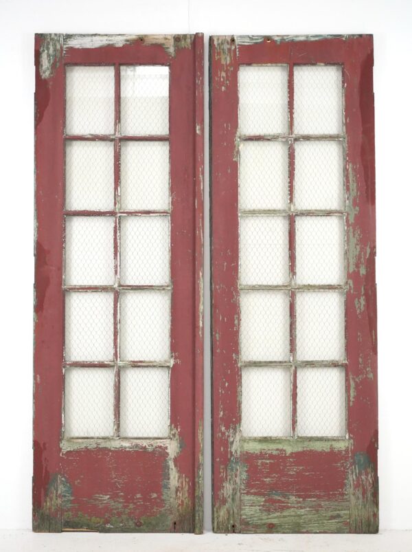 Pair of reclaimed wooden double doors with ten chicken wire glass panes each, shown with worn red paint and surface wear along the edges and bottom.