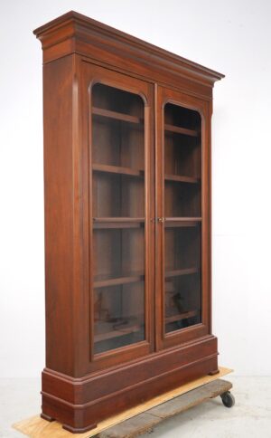 Front view of tall walnut bookcase with glass double doors, five shelves, crown molding, and wheeled wooden base.