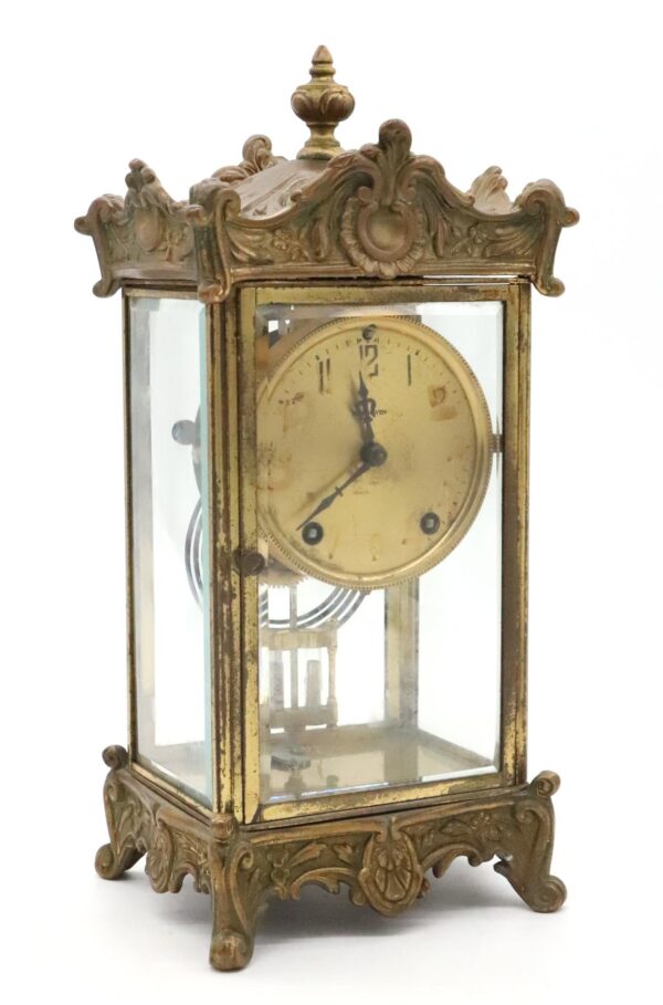 Front view highlighting the clock’s round, weathered face at 11:15 and intricate brass detailing on the wooden frame. Angled shot showing glass panels on two sides, revealing inner pendulum and gleaming brass elements. Close-up of the ornate brass corner decoration, with textured patterns visible along the wood edge. Detail of included winding key resting beside the clock base on a polished surface. Top-down view focusing on decorative molding along the upper frame and partial glass panel visibility.