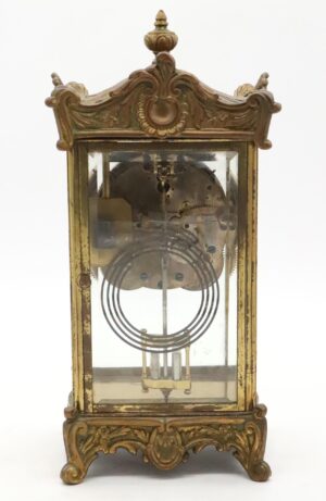 Front view of the clock showing brass bezel, glass door, and visible inner gears with ornate scrollwork framing the face. Side angle displaying decorative scrollwork on the case and revealing depth of glass panels along with a glimpse of pendulum. Close-up of exposed brass mechanism inside, highlighting detailed gearwork through open front panel. Detail shot focusing on finial at the top with wood grain texture and carved accents clearly visible. View of included key placed beside base, showing antique patina and shape designed to wind the clock mechanism.