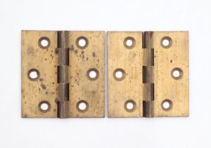 Pair of brushed brass cabinet hinges shown closed, each with five knuckles and four template holes, displaying surface wear and age marks.