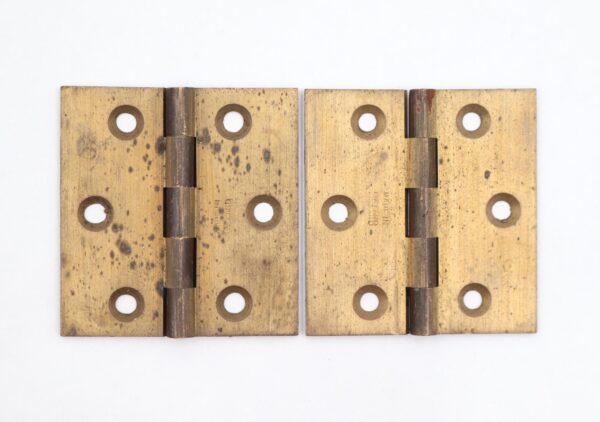 Pair of brushed brass cabinet hinges shown closed, each with five knuckles and four template holes, displaying surface wear and age marks.