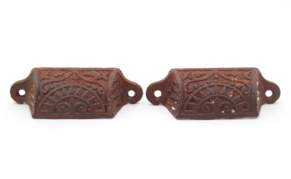 Pair of antique cast iron bin drawer pulls with an ornate Aesthetic pattern and worn black finish, shown as a matching pair with mounting holes visible on each pull.