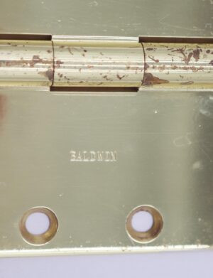 Close-up of a Baldwin polished brass butt door hinge with five knuckles and template holes, showing visible surface wear.