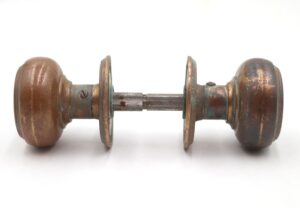 Close-up of the knobs highlighting surface rust and patina, with spindle detail visible against a white background. Angled top-down view showing both knobs’ rounded tops and connecting spindle, emphasizing weathered texture. Side view focusing on one knob’s profile and worn detailing, spindle partially obscured on a white background. Knobs displayed side by side with spindle separated, illustrating connection points and aged brass finish.