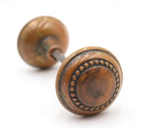Pair of antique brass passage door knobs shown with a spindle, one knob in sharp focus with a beaded edge and surface wear, and the second knob blurred behind it.