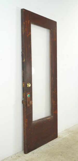 Reclaimed wood entry door with a full clear pane, shown at an angle with original brass knob set and surface wear visible.