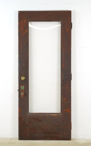 Reclaimed wood entry door with a full clear pane, dark stained brown side, and attached brass knob set and steel hinges, shown with visible surface wear and rust.