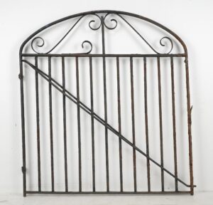 Antique wrought iron garden gate with an arched top, decorative scrollwork, and vertical bars, shown with a diagonal brace and visible surface rust and wear.