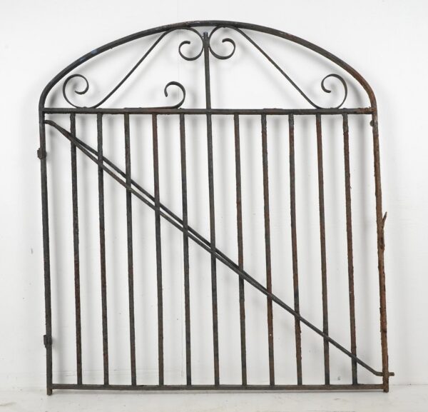 Antique wrought iron garden gate with an arched top, decorative scrollwork, and vertical bars, shown with a diagonal brace and visible surface rust and wear.