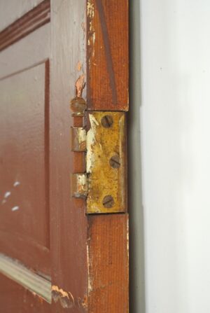 Close-up of a vintage wood passage door showing the hinge edge, with worn paint and an antique brass hinge plate visible.
