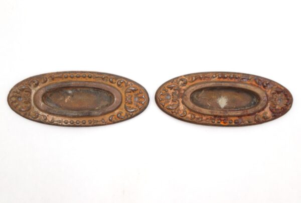 Pair of Victorian oval steel window sash lifts shown side by side, each with a recessed center and ornate border detail, with visible rust and surface wear from prior use.