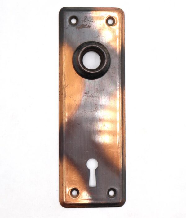 Vintage steel passage door back plate with a rectangular shape, four screw holes, a keyhole cutout, and a central circular opening, shown with a worn black and copper finish.