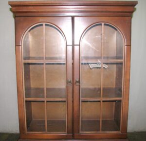 Traditional walnut hutch with arched glass doors and brass hardware, shown closed with a plastic hanger visible inside the upper cabinet.