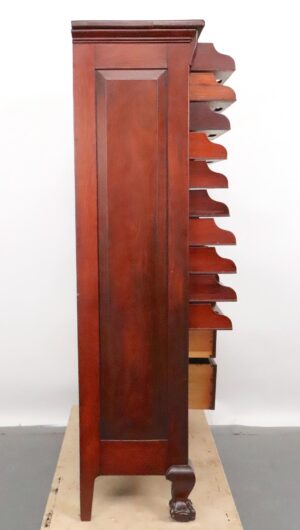 Side view of a mahogany cabinet with stacked horizontal cubbies and lower drawers, showing the tall paneled side and carved foot.