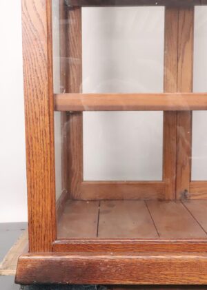 Close-up of an oak display cabinet showing the lower front corner, glass panel, and two wooden shelves.