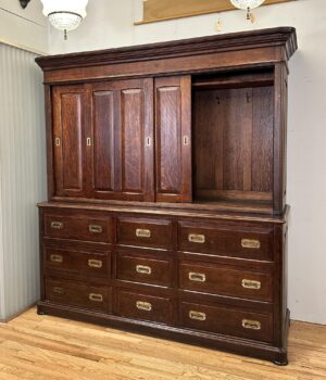 Large oak cabinet with sliding doors open to the right, showing interior hooks above nine drawers with polished brass pulls.