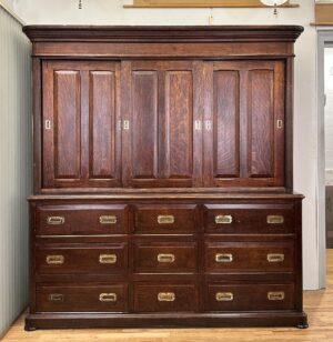 Tall dark wood cabinet with sliding top doors, nine drawers below, and recessed brass hardware.