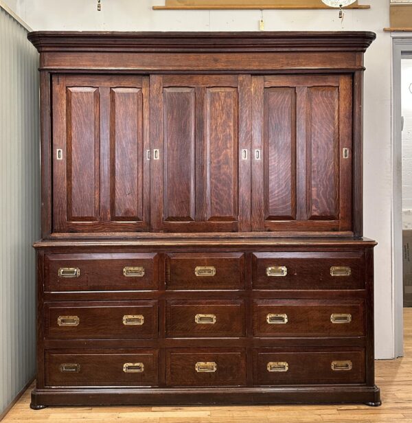 Tall dark wood cabinet with sliding top doors, nine drawers below, and recessed brass hardware.